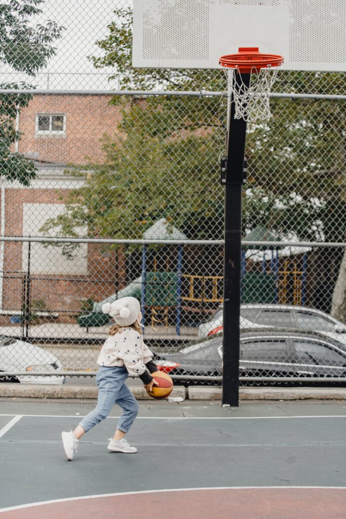 Kleines Mädchen mit Mütze spielt Basketball auf einem Freiplatz